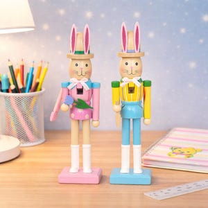 Puede incluir: Dos cascanueces de madera con forma de conejito de Pascua. Uno es rosa y el otro amarillo. Ambos tienen orejas de conejo, sombreros y botas blancas. El conejito rosa sostiene una flor azul y el conejito amarillo sostiene un cuchillo.