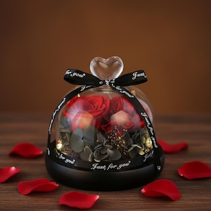 Puede incluir: Una cúpula de cristal con base negra y parte superior en forma de corazón, que alberga rosas rojas y vegetación. Una cinta negra con "Just for you" envuelve la cúpula. Pétalos de rosa rojos esparcidos.