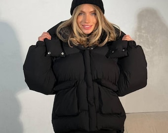 May include: A black puffer jacket with a matching black beanie. The jacket has large pockets and a high collar. The model is wearing black leggings. The outfit is a stylish winter ensemble.