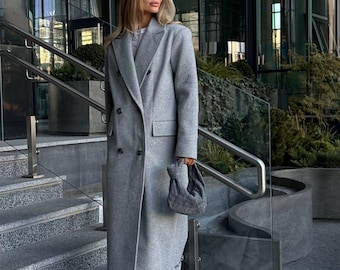 May include: A woman wearing a long, gray double-breasted coat, matching trousers, and sunglasses, descending a set of outdoor stairs. She carries a small, gray handbag. The background features a modern building with glass windows and greenery.