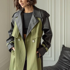 Könnte beinhalten: Ein langer olivgrüner Trenchcoat mit schwarzen Kunstlederakzenten an Schultern und Ärmeln. Der Mantel hat ein doppelreihiges Design mit zwei sichtbaren Knöpfen und einem passenden Gürtel. Das Model trägt ein schwarzes Oberteil.