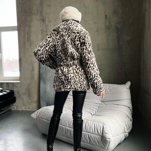 Puede incluir: Abrigo de piel sint&eacute;tica con estampado de leopardo en tonos marr&oacute;n, negro y crema. El abrigo est&aacute; ce&ntilde;ido a la cintura y se lleva con pantalones de cuero negros y botas de tac&oacute;n alto. Un sombrero de piel blanco completa el look.