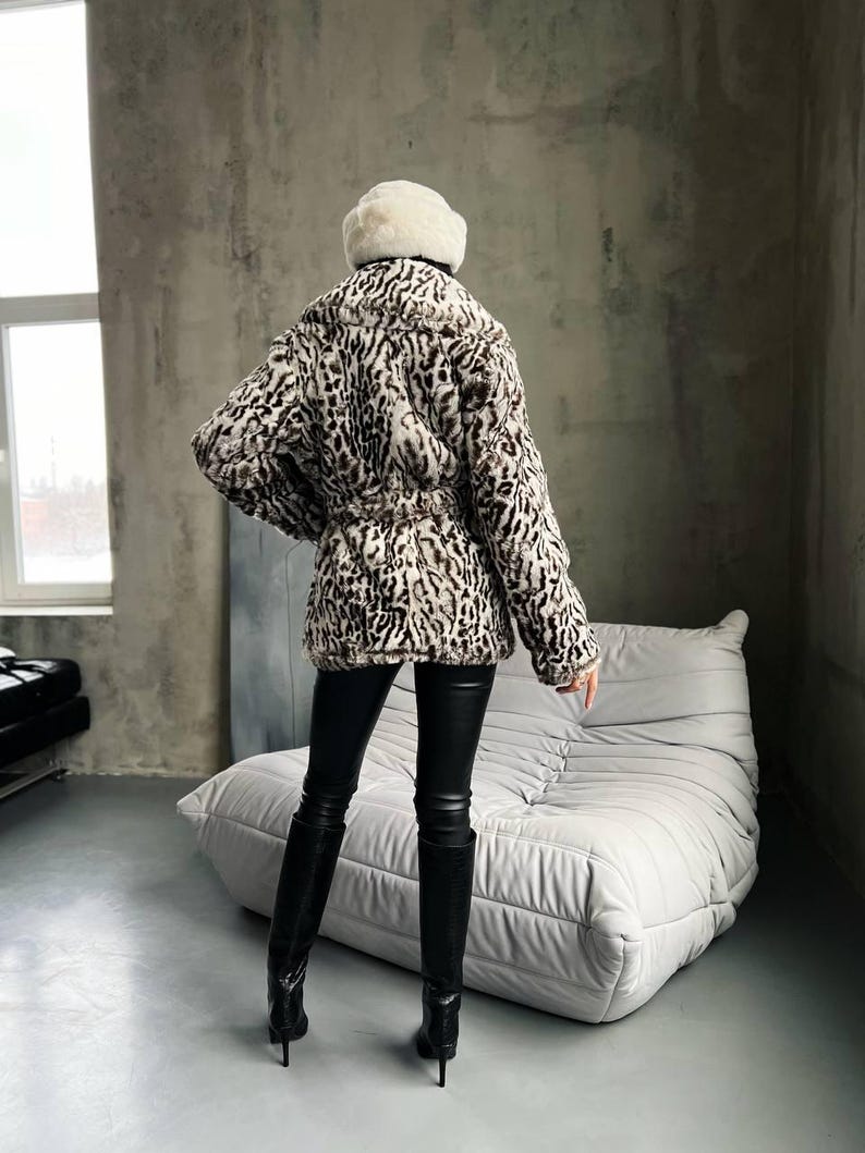 Puede incluir: Abrigo de piel sint&eacute;tica con estampado de leopardo en tonos marr&oacute;n, negro y crema. El abrigo est&aacute; ce&ntilde;ido a la cintura y se lleva con pantalones de cuero negros y botas de tac&oacute;n alto. Un sombrero de piel blanco completa el look.