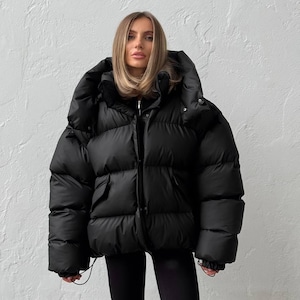 May include: A black puffer jacket with a large hood. The jacket has a zip closure and pockets. The model is wearing black leggings. The jacket appears to be made of a water-resistant material.