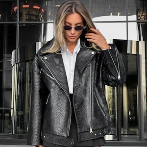 May include: A black faux leather jacket with a snakeskin texture, featuring a classic biker style with a notched collar, asymmetrical zipper, and multiple pockets. The jacket is worn over a white collared shirt and black skirt. The image also shows the text "SHOP SMAR".