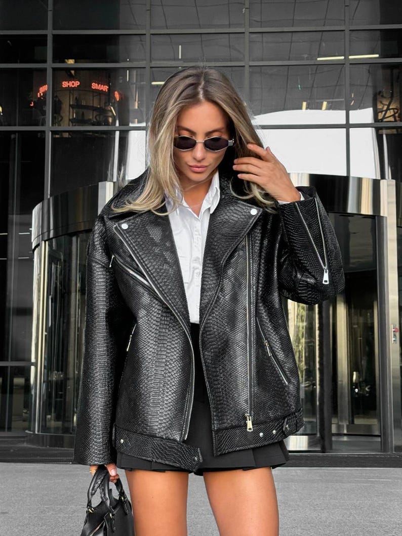 Peut inclure: Veste noire en similicuir avec une texture peau de serpent, de style motard classique avec col crant&eacute;, fermeture &eacute;clair asym&eacute;trique et plusieurs poches. La veste est port&eacute;e sur une chemise blanche et une jupe noire. Le texte "SHOP SMAR" est &eacute;galement visible.