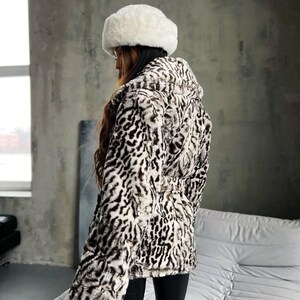 Puede incluir: Una chaqueta de piel sint&eacute;tica con capucha y un sombrero de piel blanco a juego. La chaqueta presenta un estampado de manchas blancas y negras. El modelo lleva pantalones negros. La chaqueta tiene la cintura ce&ntilde;ida.