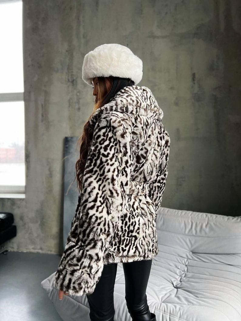 Puede incluir: Una chaqueta de piel sint&eacute;tica con capucha y un sombrero de piel blanco a juego. La chaqueta presenta un estampado de manchas blancas y negras. El modelo lleva pantalones negros. La chaqueta tiene la cintura ce&ntilde;ida.