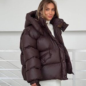 May include: A dark brown puffer jacket with a large hood and pockets. The jacket is worn over a white sweater and skirt. The jacket has a puffy, quilted design and a zipper closure. The model is holding a dark brown tote bag.