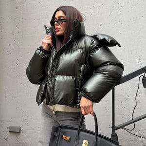 Könnte beinhalten: Eine schwarze Steppjacke mit hohem Kragen und Reißverschluss. Die Jacke wird mit grauen Jeans und einer schwarzen Lederhandtasche mit goldenen Details kombiniert. Die Person trägt eine Sonnenbrille.