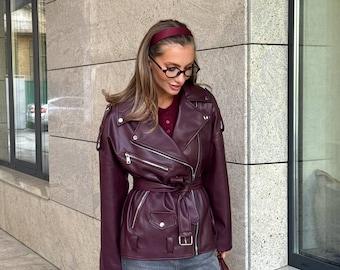 Könnte beinhalten: Burgunderfarbene Kunstlederjacke mit Gürtel in der Taille und silbernen Reißverschlüssen. Die Jacke hat einen klassischen Biker-Stil mit Reverskragen und Schulterklappen. Das Model trägt eine graue Jeans und ein bordeauxfarbenes Stirnband.