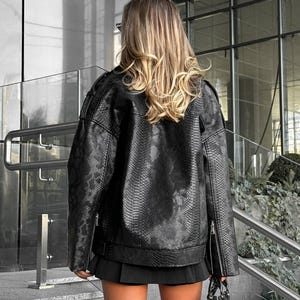 Peut inclure: Veste noire en similicuir avec une texture peau de serpent, fermeture &eacute;clair et petit sac &agrave; main noir. La veste a un style motard classique avec une coupe courte et carr&eacute;e. La mod&egrave;le porte une jupe noire.