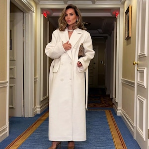 May include: A long, white double-breasted coat with a matching belt. The coat is worn with brown heels and a brown clutch. The hallway has blue carpet with a gold border and white walls. The coat's length is approximately 150 cm.