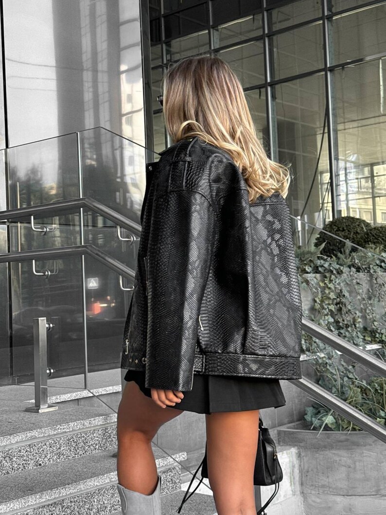 Peut inclure: Veste noire en similicuir avec un motif peau de serpent. La veste a un style motard classique avec un col crant&eacute;, des d&eacute;tails de fermeture &eacute;clair et une coupe l&eacute;g&egrave;rement courte. La mod&egrave;le porte une jupe noire et des bottes grises.