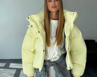 May include: A light yellow puffer jacket with a high collar and button closure. The jacket is worn over a white t-shirt and a blue plaid shirt tied around the waist. The model has long blonde hair.
