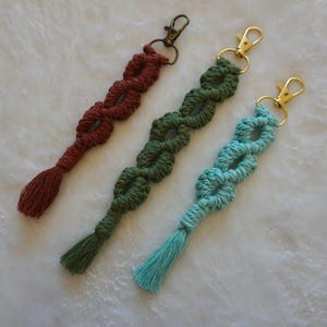 May include: Three macrame keychains in shades of red, green, and blue. Each keychain features a knotted design with a tassel at the end and a metal clasp for attachment. The keychains are displayed on a white, textured surface.
