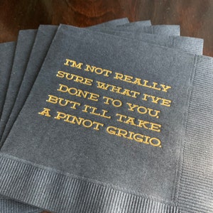 May include: A stack of navy blue cocktail napkins with gold lettering. The text reads: "I'M NOT REALLY SURE WHAT I'VE DONE TO YOU, BUT I'LL TAKE A PINOT GRIGIO."