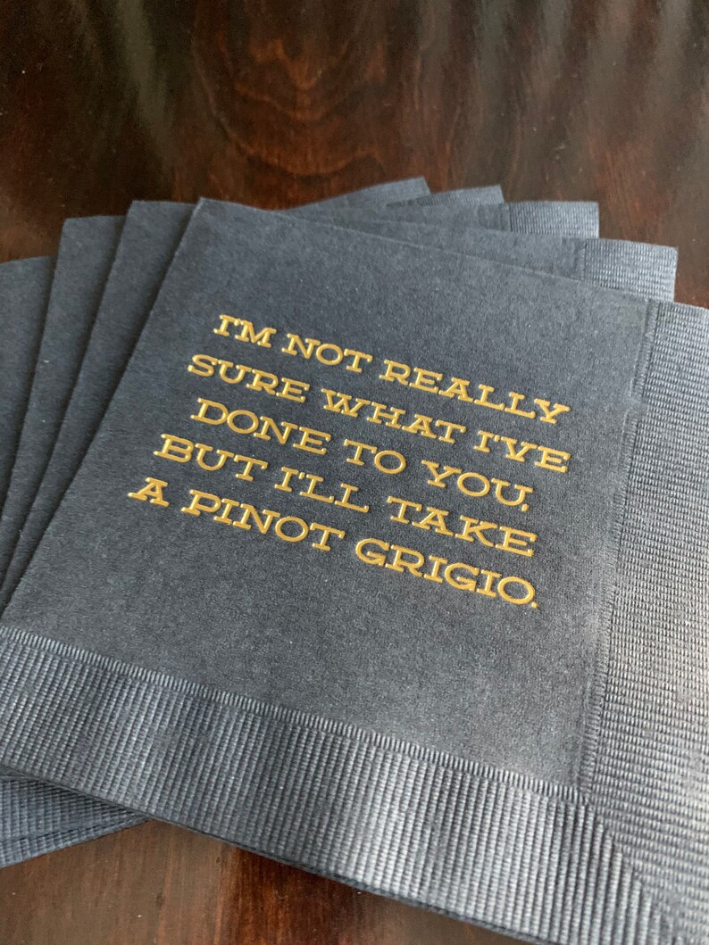 May include: A stack of navy blue cocktail napkins with gold lettering. The text reads: "I'M NOT REALLY SURE WHAT I'VE DONE TO YOU, BUT I'LL TAKE A PINOT GRIGIO."