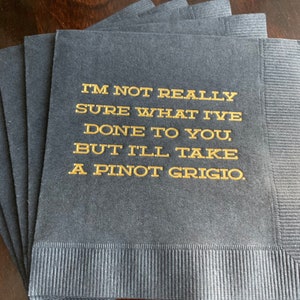 May include: A stack of navy blue cocktail napkins with gold lettering. The text reads, "I'M NOT REALLY SURE WHAT I'VE DONE TO YOU, BUT I'LL TAKE A PINOT GRIGIO."