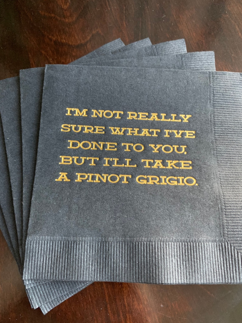 May include: A stack of navy blue cocktail napkins with gold lettering. The text reads, "I'M NOT REALLY SURE WHAT I'VE DONE TO YOU, BUT I'LL TAKE A PINOT GRIGIO."