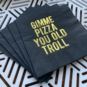 May include: A stack of black napkins with gold foil lettering that reads "Gimme Pizza You Old Troll".