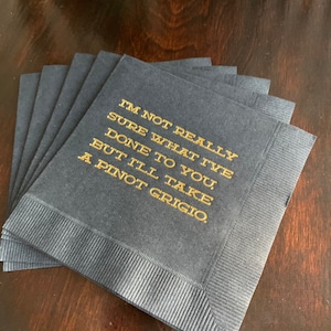 May include: A stack of navy blue cocktail napkins with gold text that reads "I'M NOT REALLY SURE WHAT I'VE DONE TO YOU, BUT I'LL TAKE A PINOT GRIGIO."