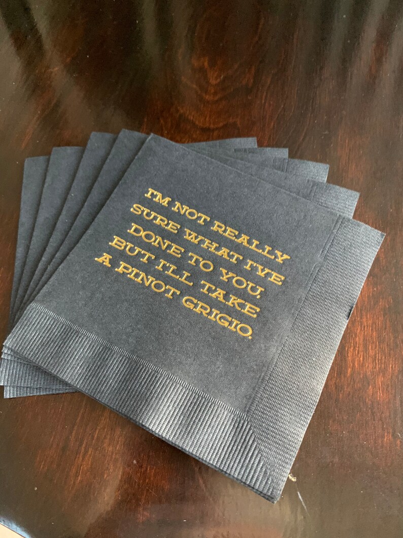 May include: A stack of navy blue cocktail napkins with gold text that reads "I'M NOT REALLY SURE WHAT I'VE DONE TO YOU, BUT I'LL TAKE A PINOT GRIGIO."