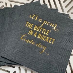 May include: A stack of black napkins with gold foil lettering that reads "it's a pour the bottle in a bucket kinda day".
