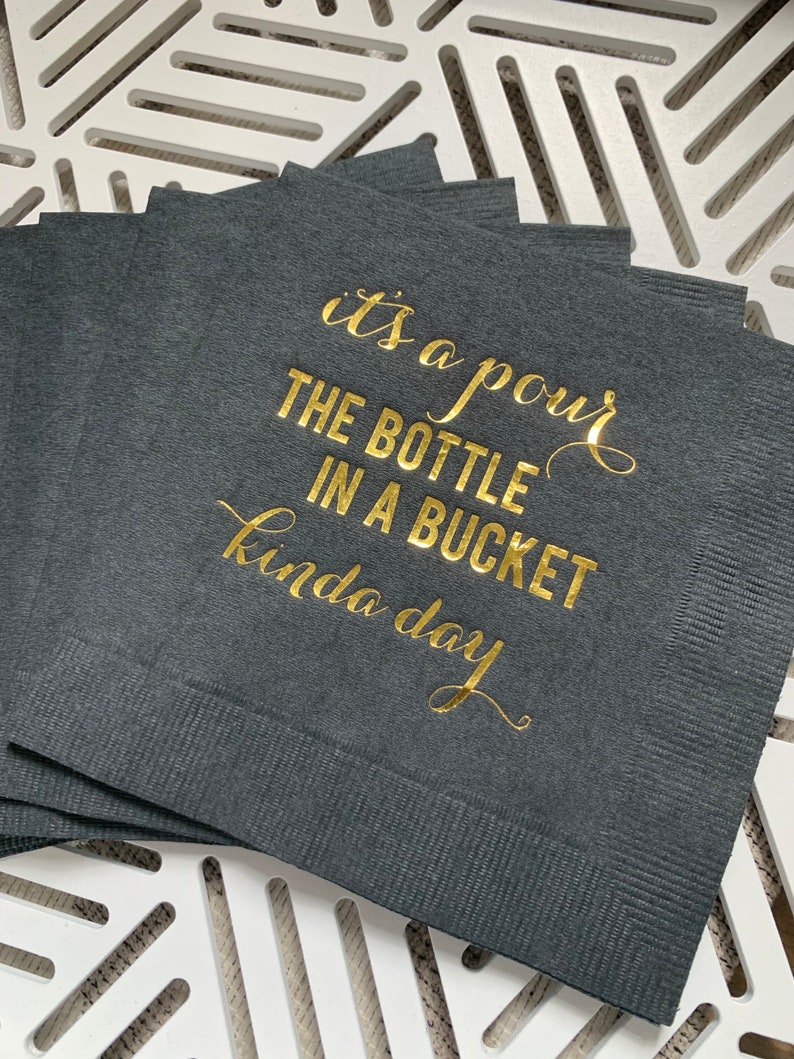 May include: A stack of black napkins with gold foil lettering that reads "it's a pour the bottle in a bucket kinda day".