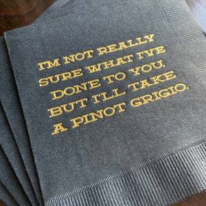 May include: A stack of dark gray cocktail napkins with gold lettering. The text reads "I'M NOT REALLY SURE WHAT I'VE DONE TO YOU, BUT I'LL TAKE A PINOT GRIGIO."