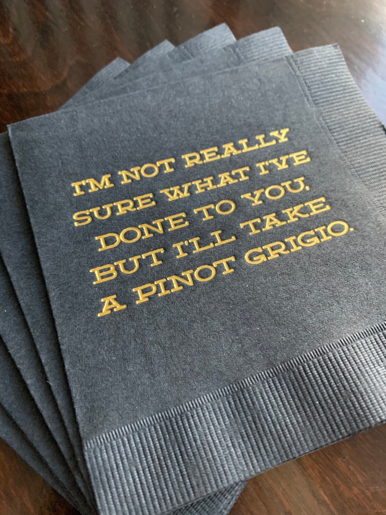 May include: A stack of dark gray cocktail napkins with gold lettering. The text reads "I'M NOT REALLY SURE WHAT I'VE DONE TO YOU, BUT I'LL TAKE A PINOT GRIGIO."