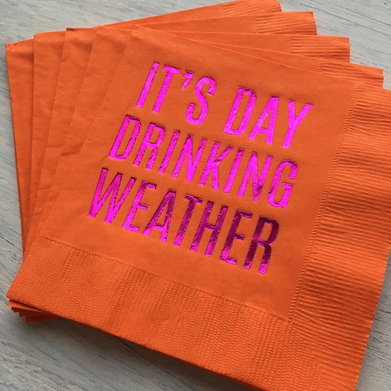 May include: A stack of orange paper napkins with the text "It's Day Drinking Weather" printed in pink foil.
