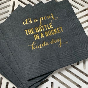 May include: A stack of black napkins with gold foil lettering that reads "it's a pour the bottle in a bucket kinda day".