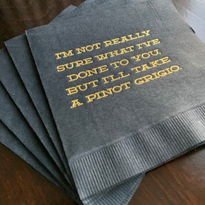 May include: A stack of black cocktail napkins with gold lettering. The text reads: "I'M NOT REALLY SURE WHAT I'VE DONE TO YOU, BUT I'LL TAKE A PINOT GRIGIO."