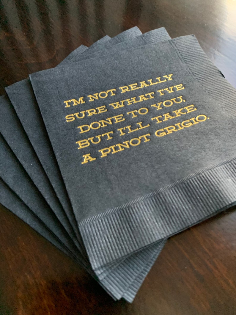 May include: A stack of black cocktail napkins with gold lettering. The text reads: "I'M NOT REALLY SURE WHAT I'VE DONE TO YOU, BUT I'LL TAKE A PINOT GRIGIO."