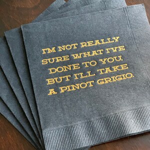 May include: A stack of dark gray cocktail napkins with gold text that reads "I'M NOT REALLY SURE WHAT I'VE DONE TO YOU, BUT I'LL TAKE A PINOT GRIGIO."