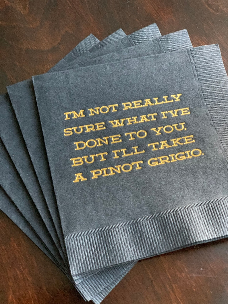 May include: A stack of dark gray cocktail napkins with gold text that reads "I'M NOT REALLY SURE WHAT I'VE DONE TO YOU, BUT I'LL TAKE A PINOT GRIGIO."