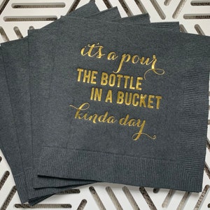 May include: A stack of dark gray paper napkins with gold foil lettering that reads "it's a pour the bottle in a bucket kinda day".