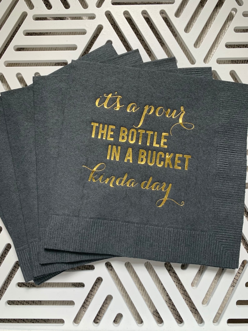 May include: A stack of dark gray paper napkins with gold foil lettering that reads "it's a pour the bottle in a bucket kinda day".