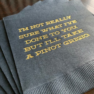 May include: A stack of dark gray cocktail napkins with gold lettering. The text reads, "I'M NOT REALLY SURE WHAT I'VE DONE TO YOU, BUT I'LL TAKE A PINOT GRIGIO."