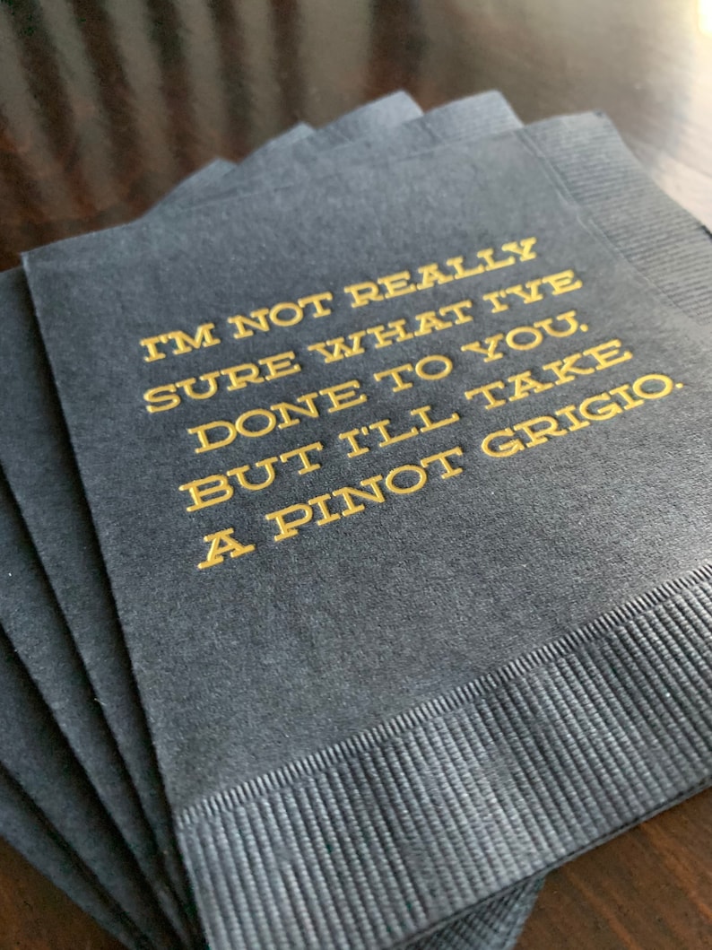 May include: A stack of dark gray cocktail napkins with gold lettering. The text reads, "I'M NOT REALLY SURE WHAT I'VE DONE TO YOU, BUT I'LL TAKE A PINOT GRIGIO."