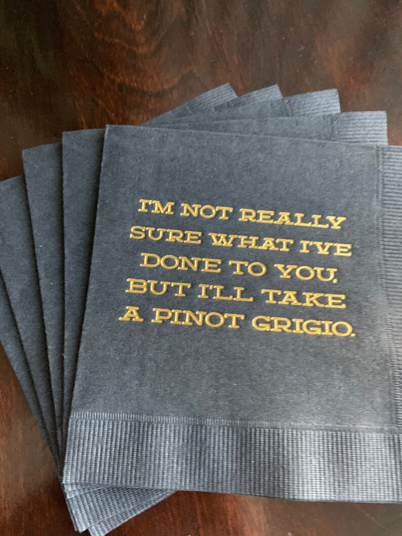 May include: A stack of navy blue cocktail napkins with gold lettering. The text reads, "I'M NOT REALLY SURE WHAT I'VE DONE TO YOU, BUT I'LL TAKE A PINOT GRIGIO."