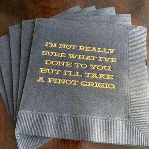 May include: A stack of navy blue paper napkins with gold lettering. The text reads, "I'M NOT REALLY SURE WHAT I'VE DONE TO YOU, BUT I'LL TAKE A PINOT GRIGIO."