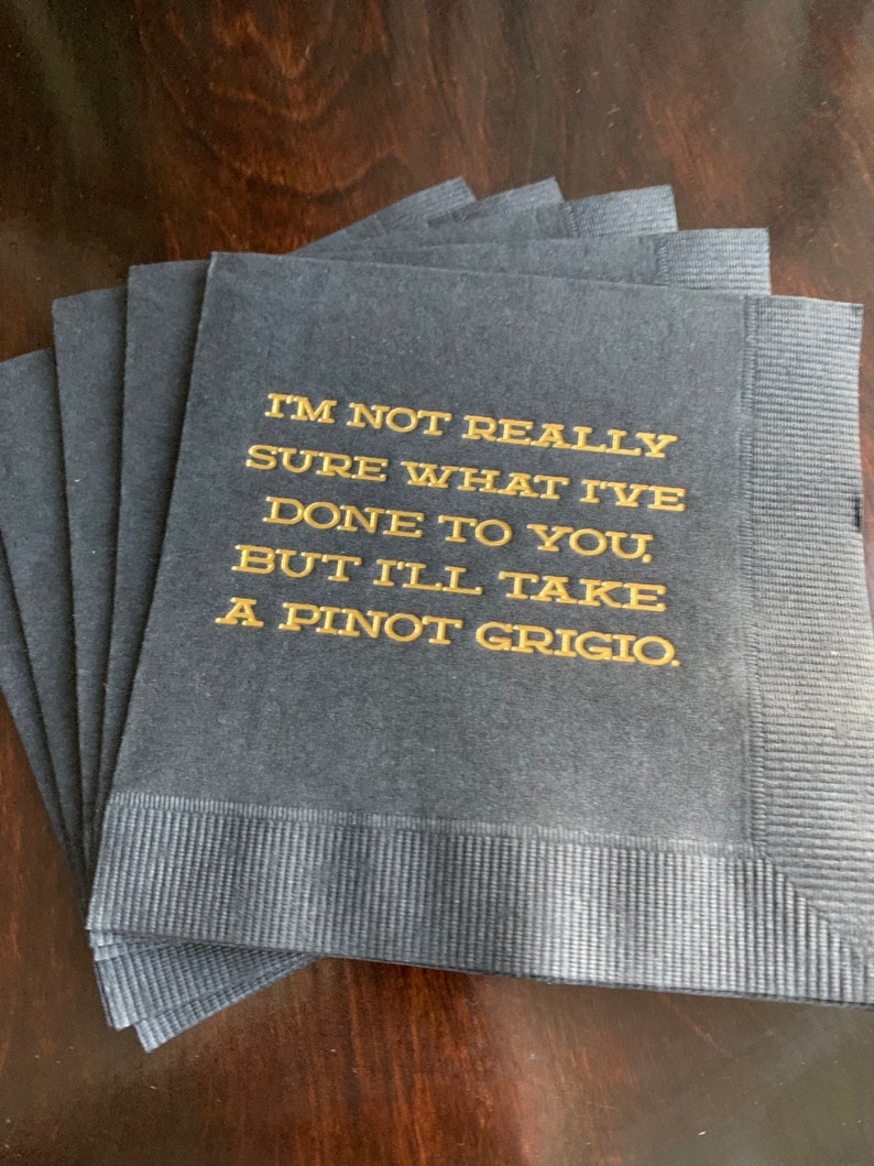 May include: A stack of navy blue paper napkins with gold lettering. The text reads, "I'M NOT REALLY SURE WHAT I'VE DONE TO YOU, BUT I'LL TAKE A PINOT GRIGIO."