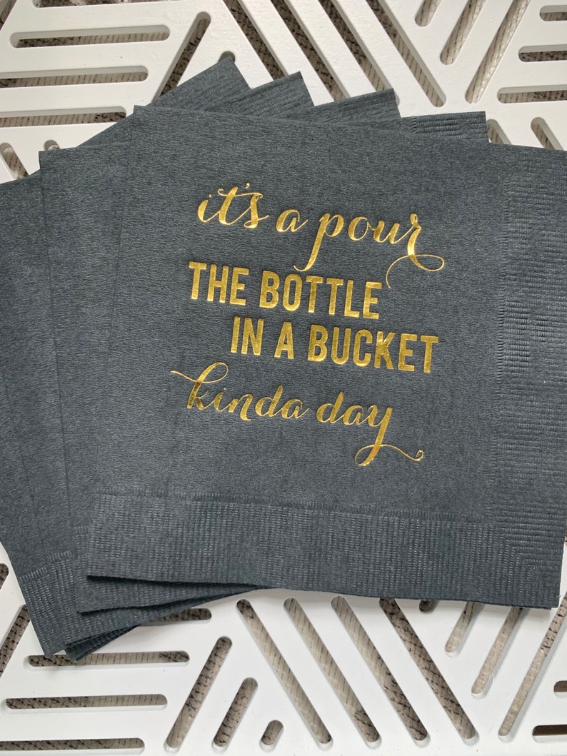 May include: A stack of dark gray napkins with gold foil lettering that reads "it's a pour the bottle in a bucket kinda day".