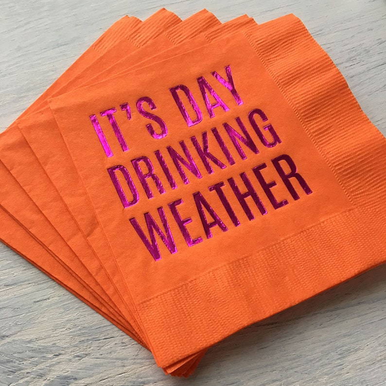 May include: A stack of orange paper napkins with the text "It's Day Drinking Weather" printed in pink foil.