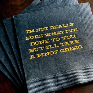 May include: A stack of dark gray cocktail napkins with gold text. The text reads: "I'M NOT REALLY SURE WHAT I'VE DONE TO YOU, BUT I'LL TAKE A PINOT GRIGIO."
