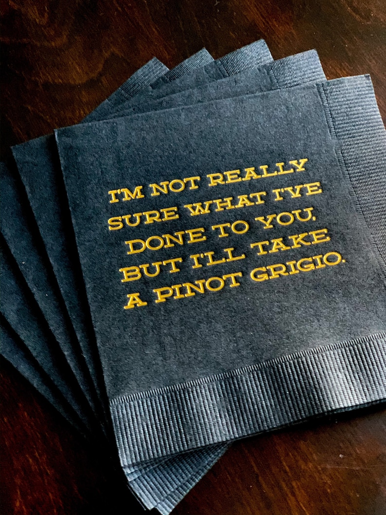 May include: A stack of dark gray cocktail napkins with gold text. The text reads: "I'M NOT REALLY SURE WHAT I'VE DONE TO YOU, BUT I'LL TAKE A PINOT GRIGIO."