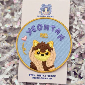 May include: A round embroidered patch featuring a cartoon dog with the name "Yeontan" in purple letters. The patch has a light blue background and a gold border. The text "ETSY | INSTA | TIKTOK @SEOULFULNOONA" is printed below the patch.