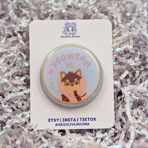 May include: A round pin with a light blue background featuring a cartoon dog with the name "Yeontan" in pink lettering. The pin is on a white card with the text "ETSY | INSTA | TIKTOK @SEOULFULNOONA".